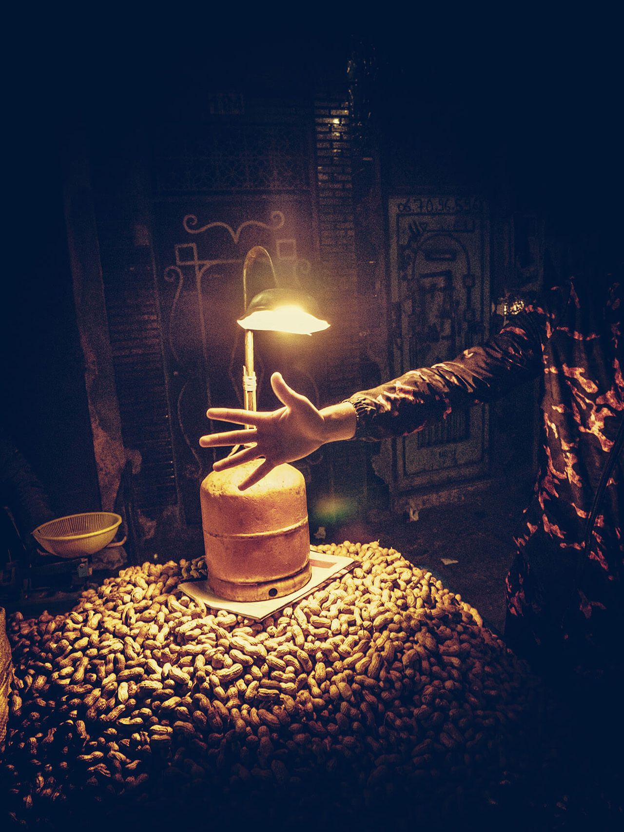 A hand reaches toward a glowing lamp above a pile of roasted nuts at a night market stall