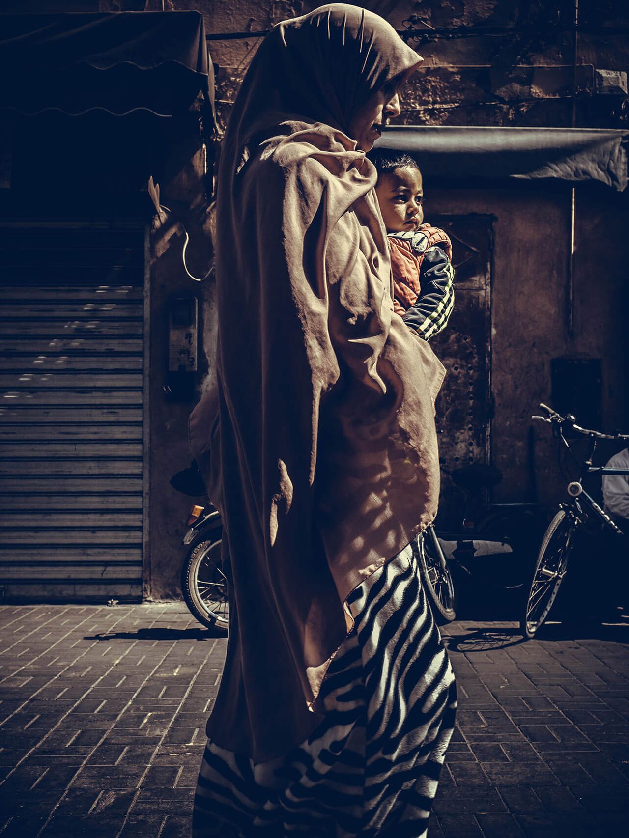 A woman in a headscarf carries a child past a shuttered storefront