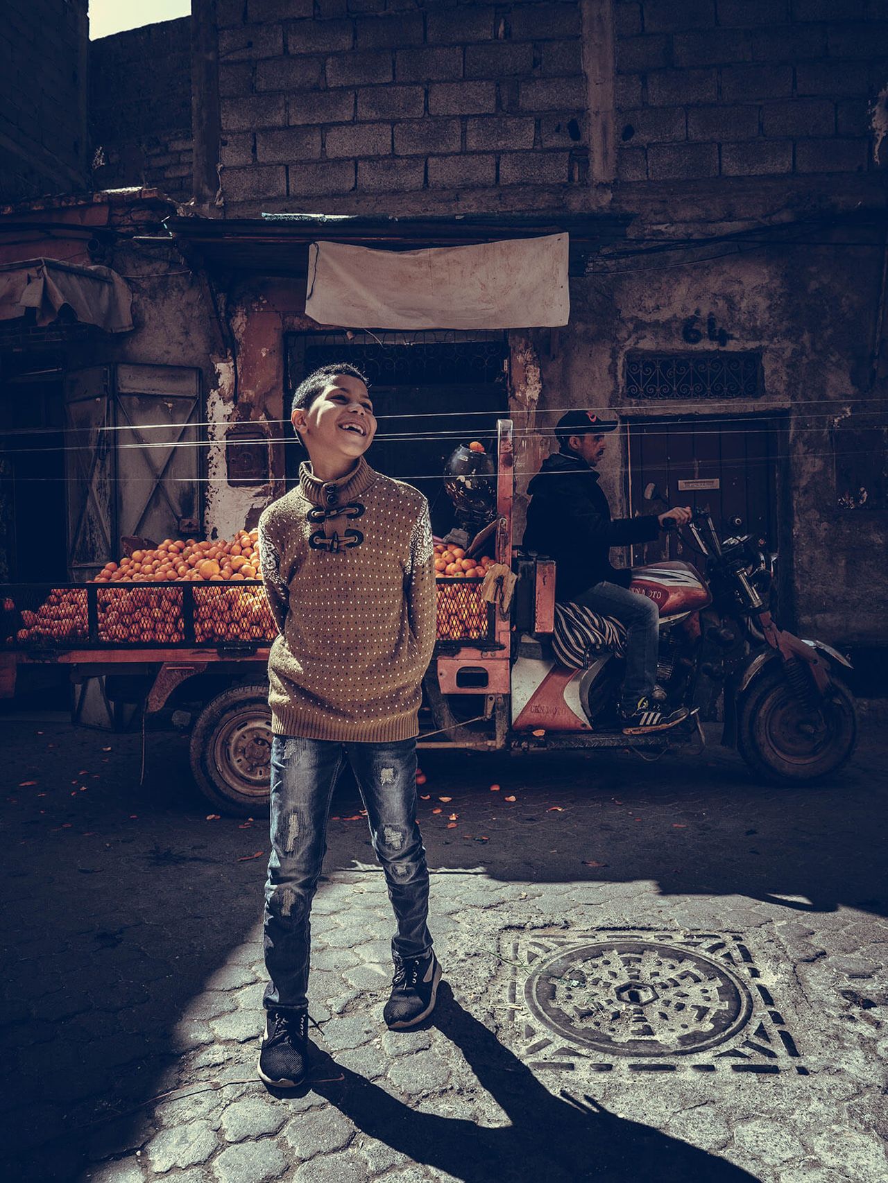 A smiling boy stands in a patch of sunlight in front of a fruit cart