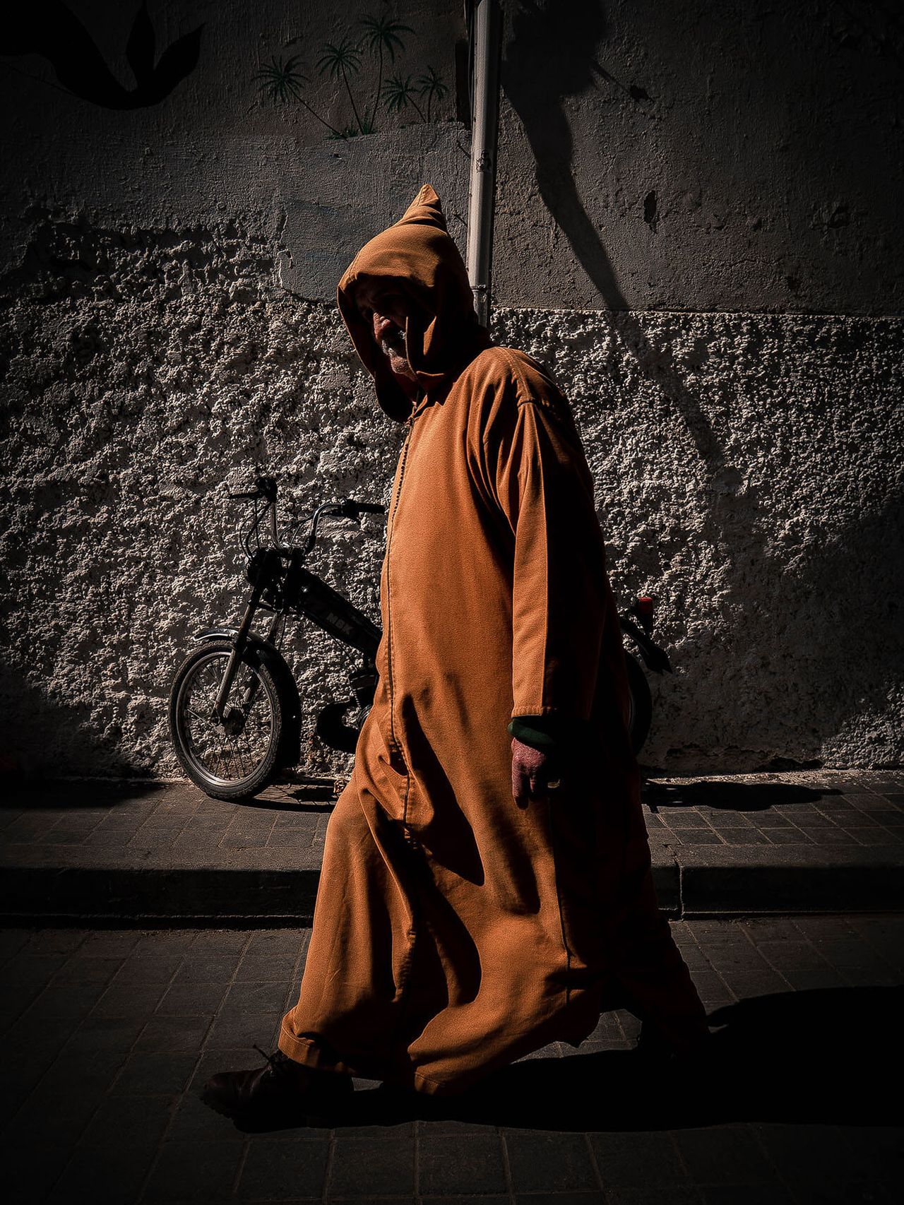 A man in a hooded orange robe walks past a bicycle leaning on a wall