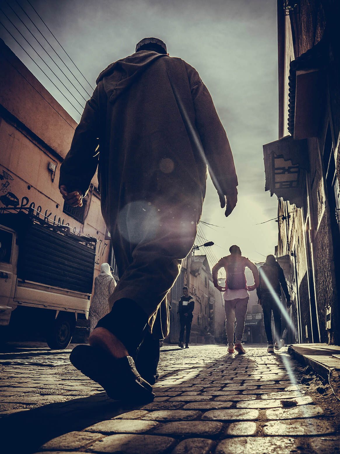 A man in a long coat strides down a sunlit cobblestone alley