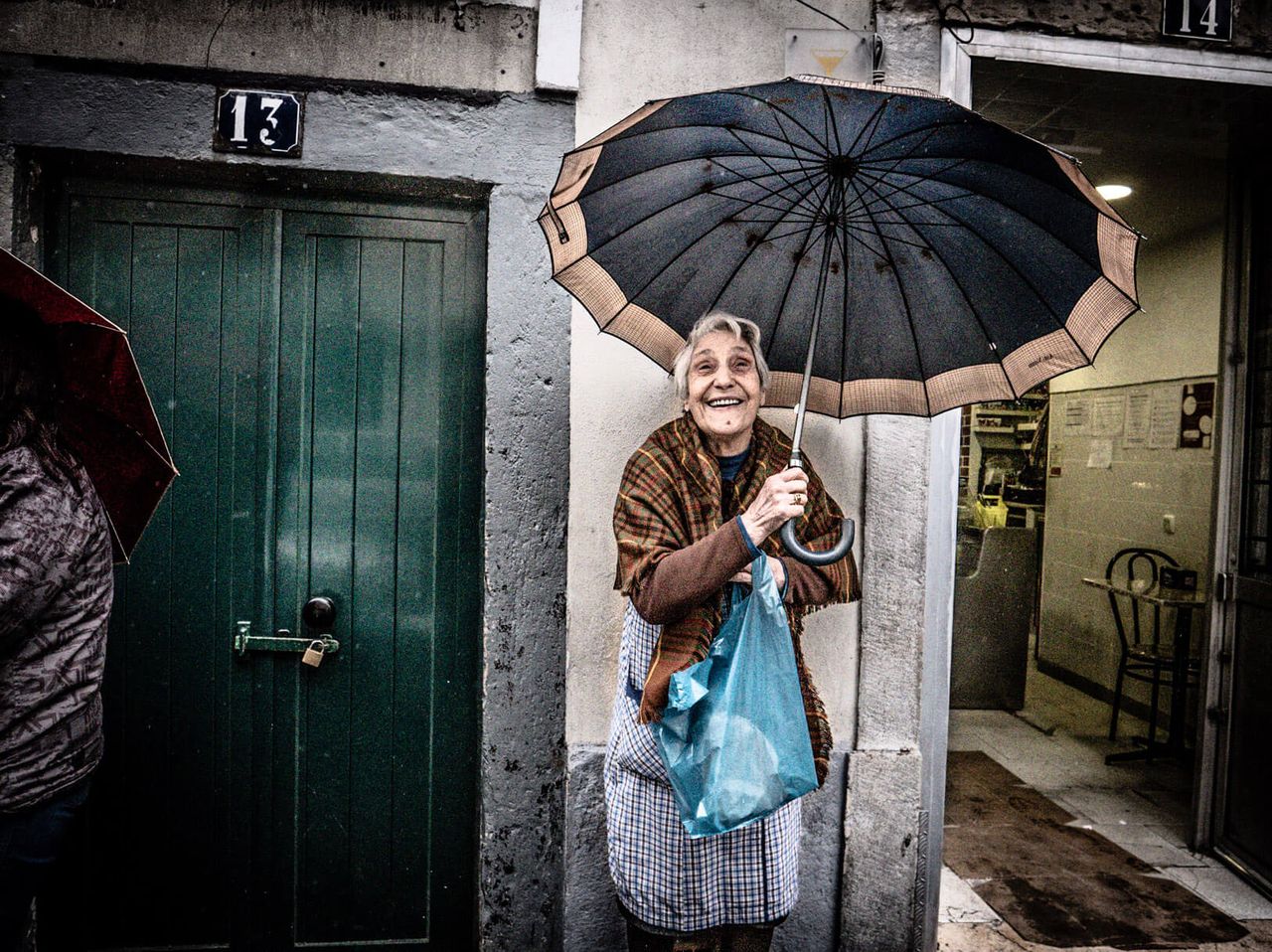 A grinning older woman holds an umbrella in a narrow doorway