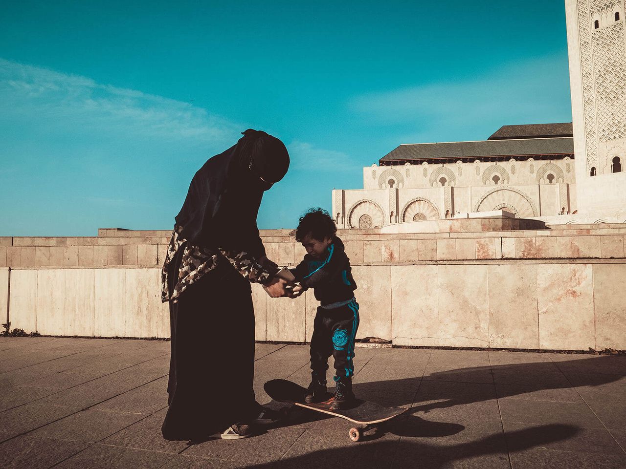 A mother steadies a child on a skateboard under a bright blue sky