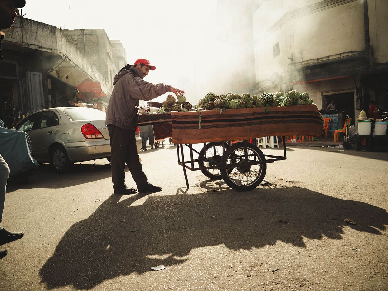 A vendor arranges produce on a wooden cart in hazy morning light